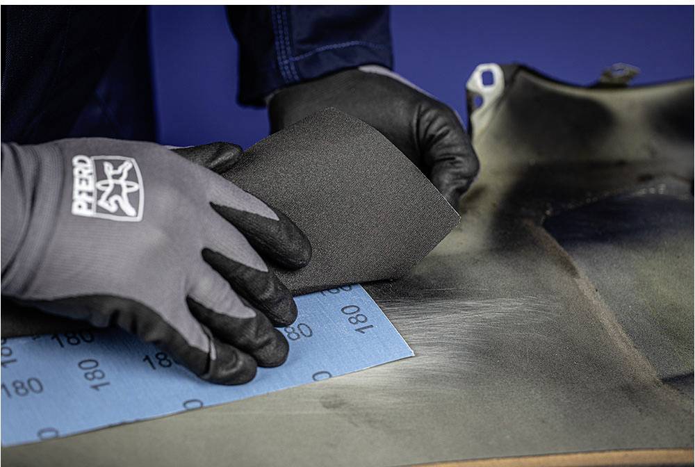 A person is wearing gloves and sanding a metal surface with sandpaper. The sandpaper has a grit of 180.