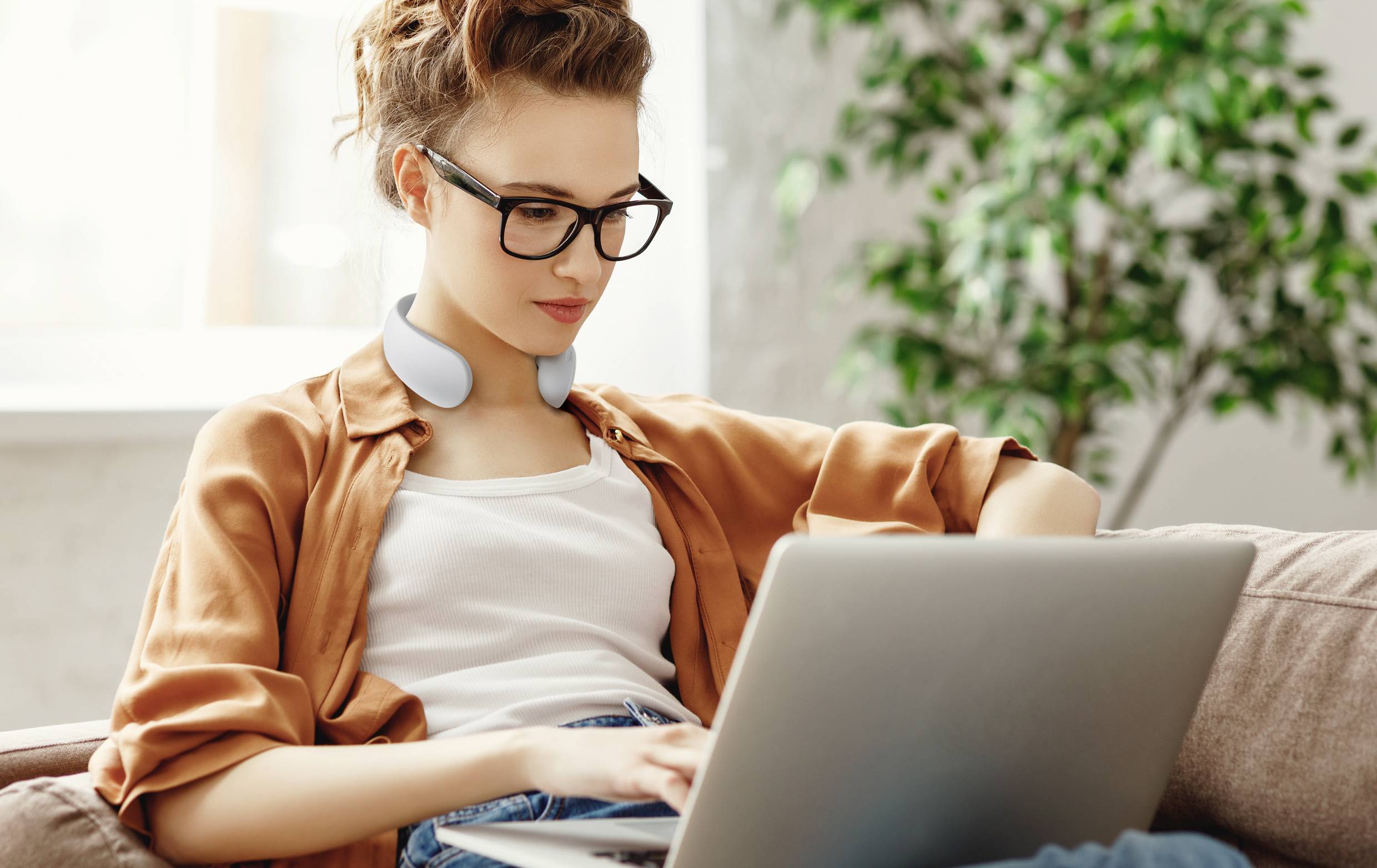 A person is sitting on a sofa, wearing headphones around their neck, and working intently on a laptop. A green plant stands in the background.
