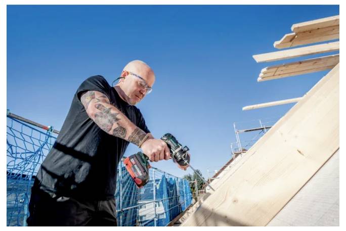 A person wearing safety glasses is using a power drill on a wooden structure at a construction site under a clear blue sky.