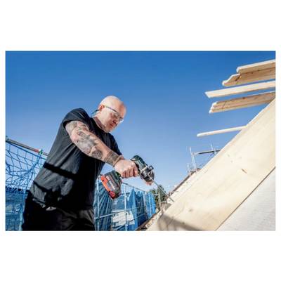 A person wearing safety glasses is using a power drill on a wooden structure at a construction site under a clear blue sky.