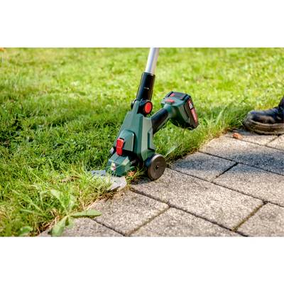 A cordless grass trimmer being used to cut grass along a stone walkway. The trimmer has a green body and appears to be battery-operated.
