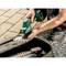 A person adjusts the blade of a green power tool, with a gravel and paved surface in the background. A case with additional tools is nearby.