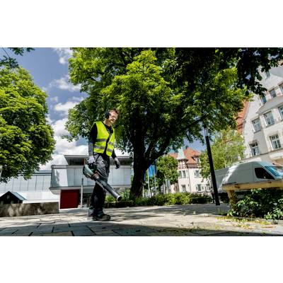 A person wearing a high-visibility yellow vest is using a leaf blower on a pavement beneath a large tree in an urban setting.