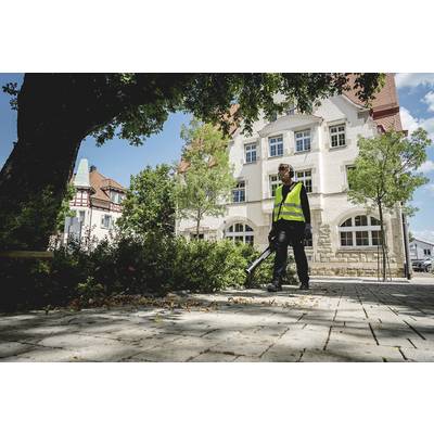 A person wearing a high-visibility yellow vest is using a leaf blower to clear leaves from a paved path. A building can be seen in the background.