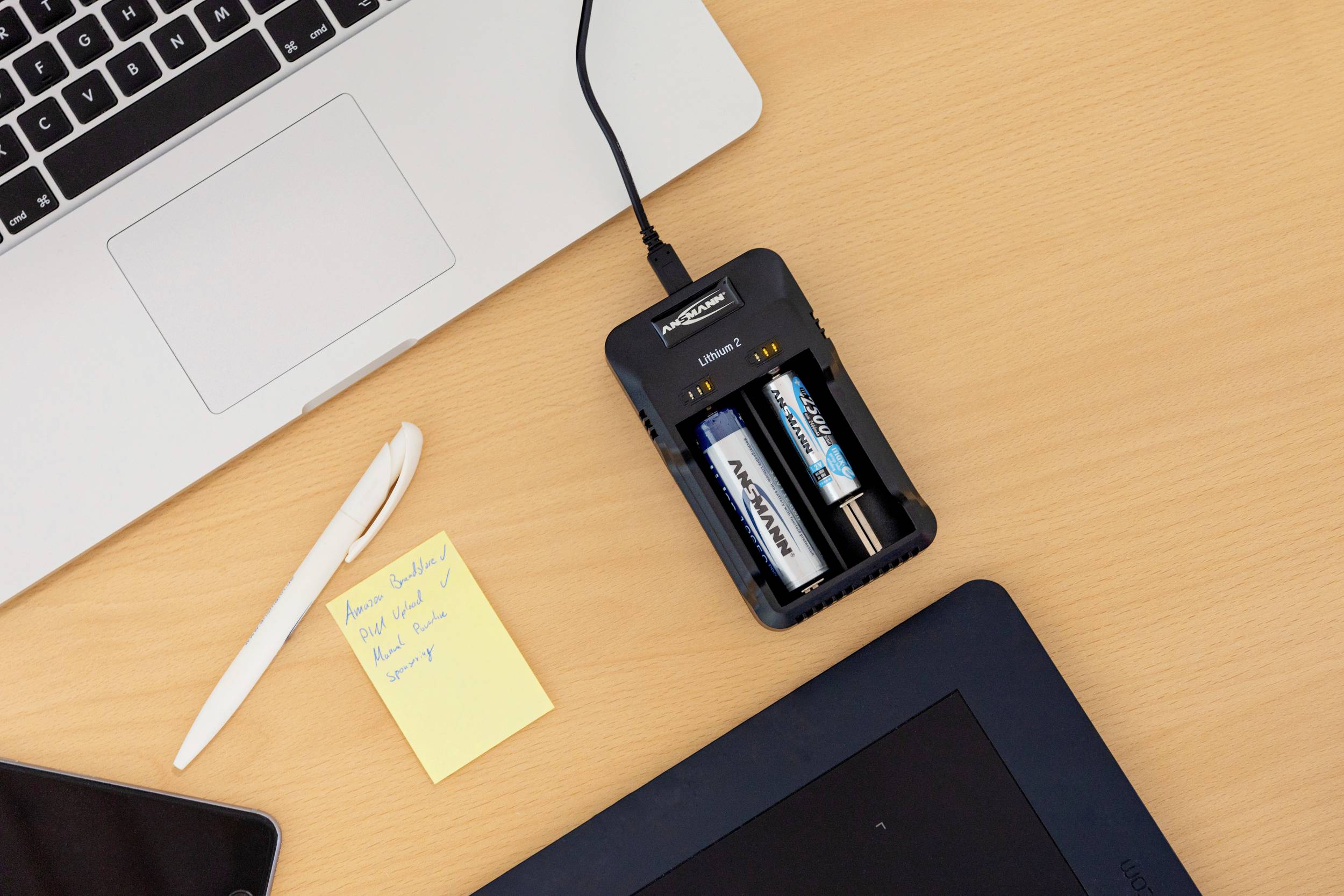 A table with a laptop, a pen, a yellow sticky note next to a battery charger with two different AA batteries.