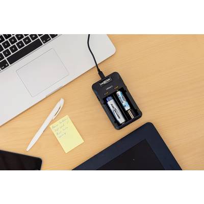 A table with a laptop, a pen, a yellow sticky note next to a battery charger with two different AA batteries.