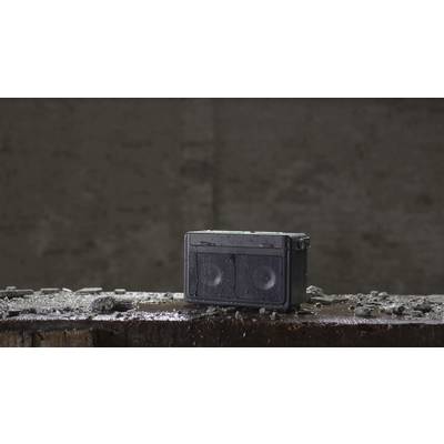A robust, waterproof speaker stands on a concrete block, surrounded by rubble, in front of a grey, textured wall.