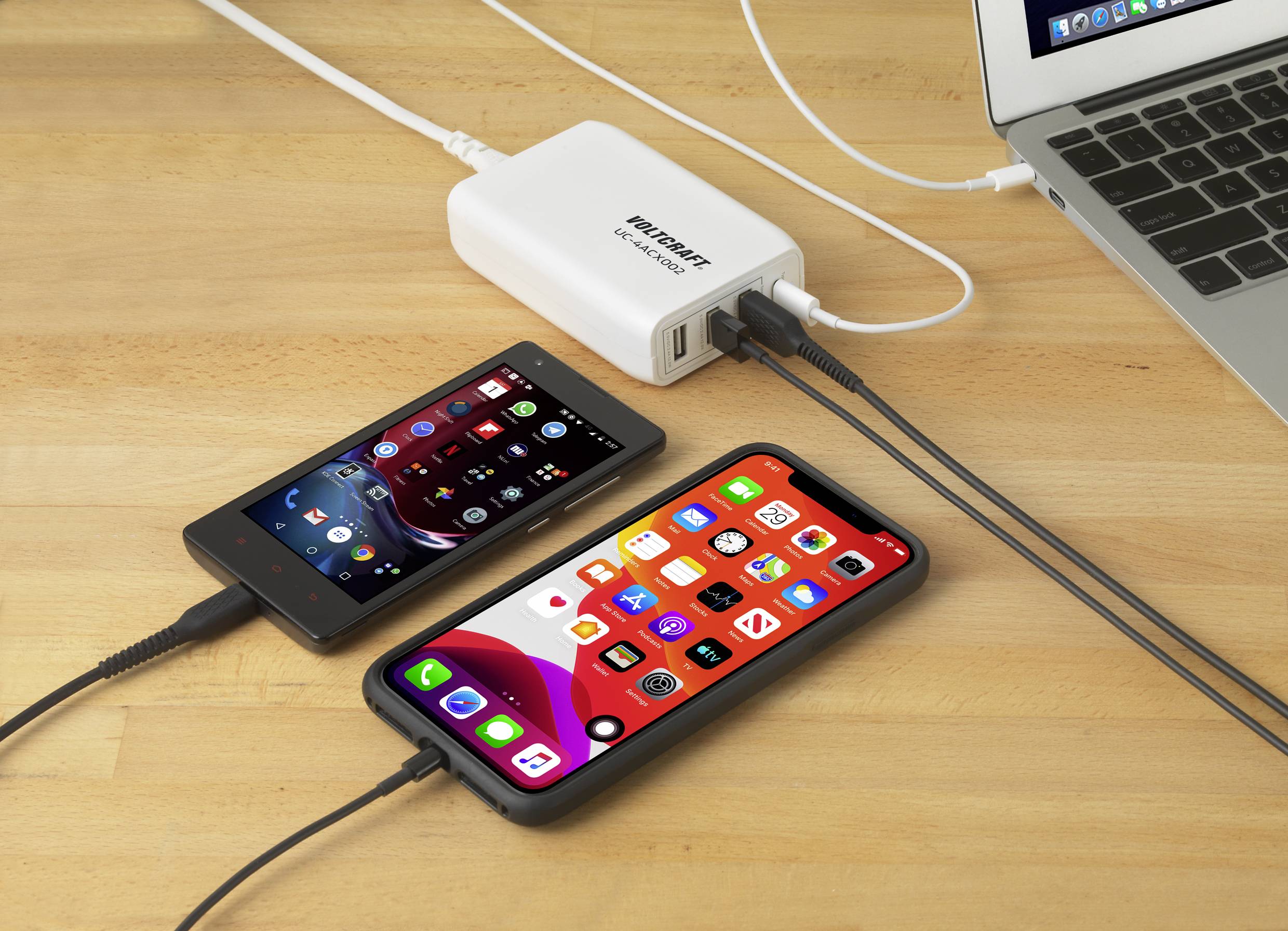 A smartphone and tablet are lying on a wooden table, connected to a laptop and a charger plugged into a power socket.