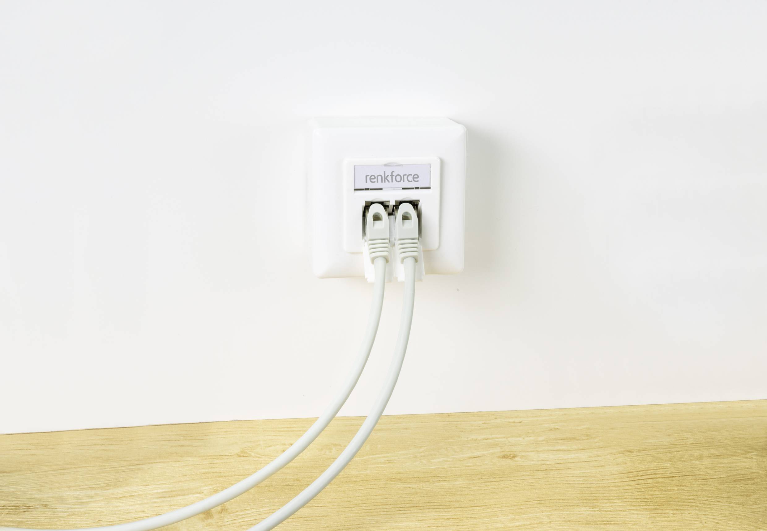 A white network wall socket with three connected Ethernet cables in front of a white wall on a wooden surface.