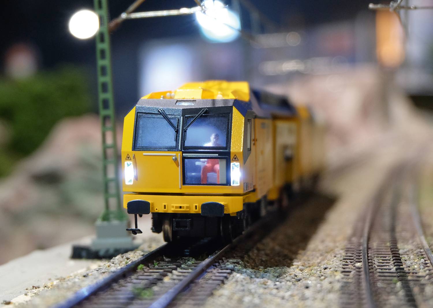 A yellow model train is travelling along a miniature railway track. The front of the train is sharply in focus, while the background is blurred.