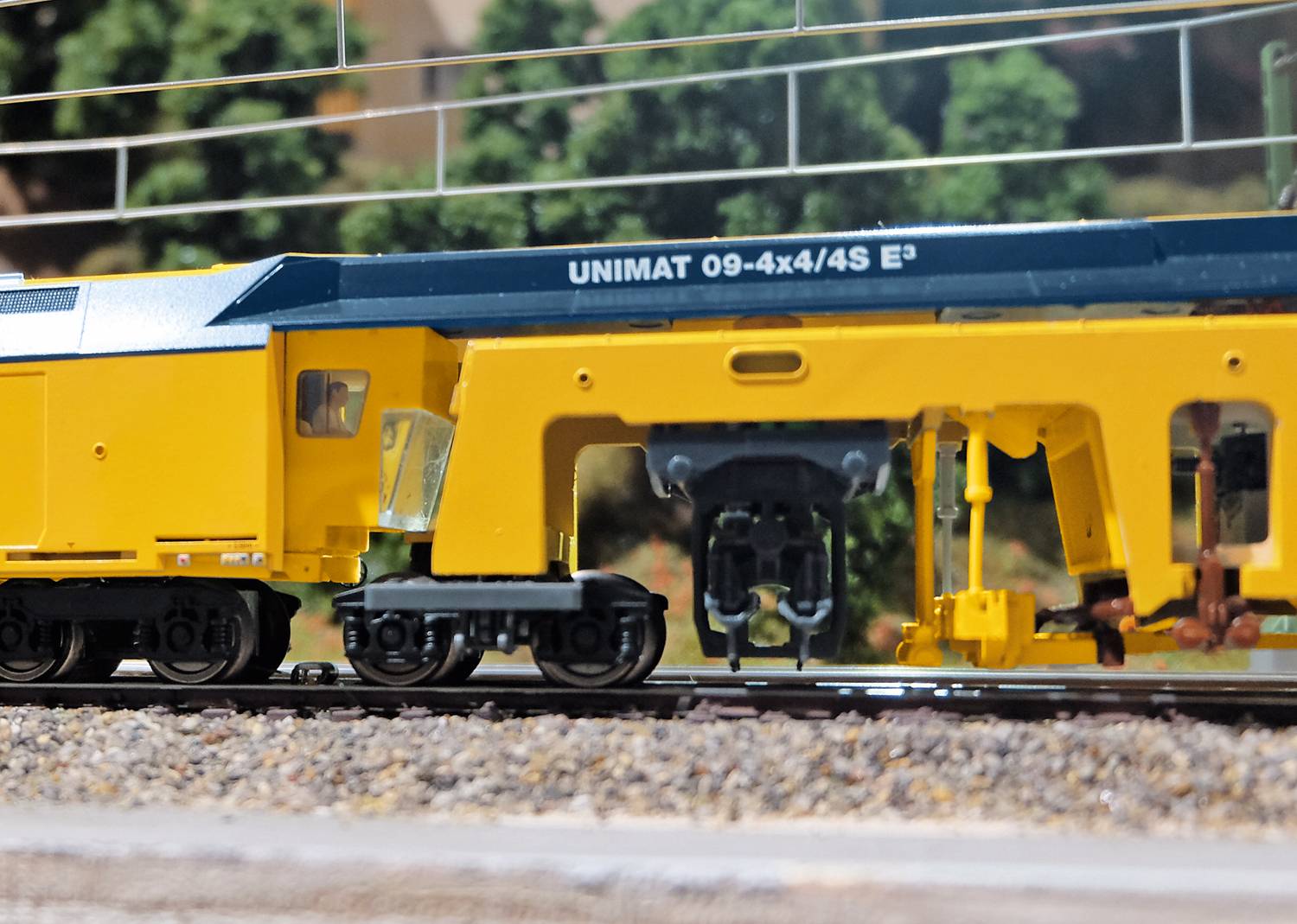 The image shows a yellow model train of the 'UNIMAT 09-4x4/4S E3' brand on a track. Blurry trees can be seen in the background.