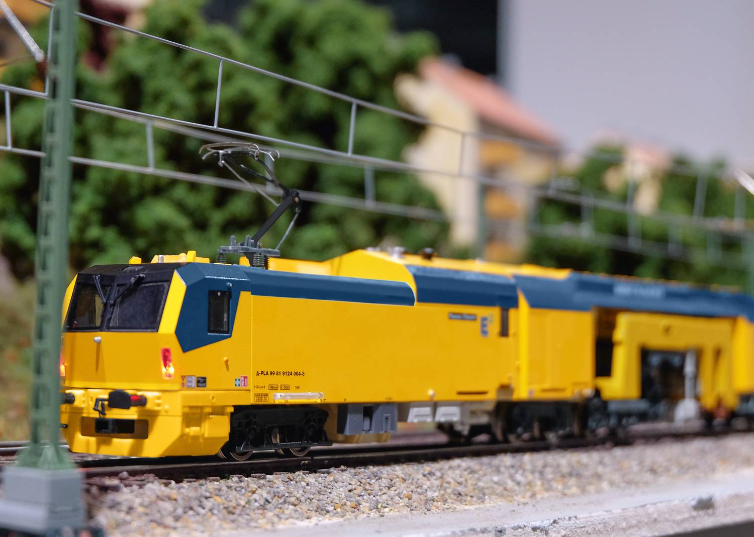 A yellow model train on a miniature railway. The train is travelling along an electric overhead line, with trees and buildings visible in the background.