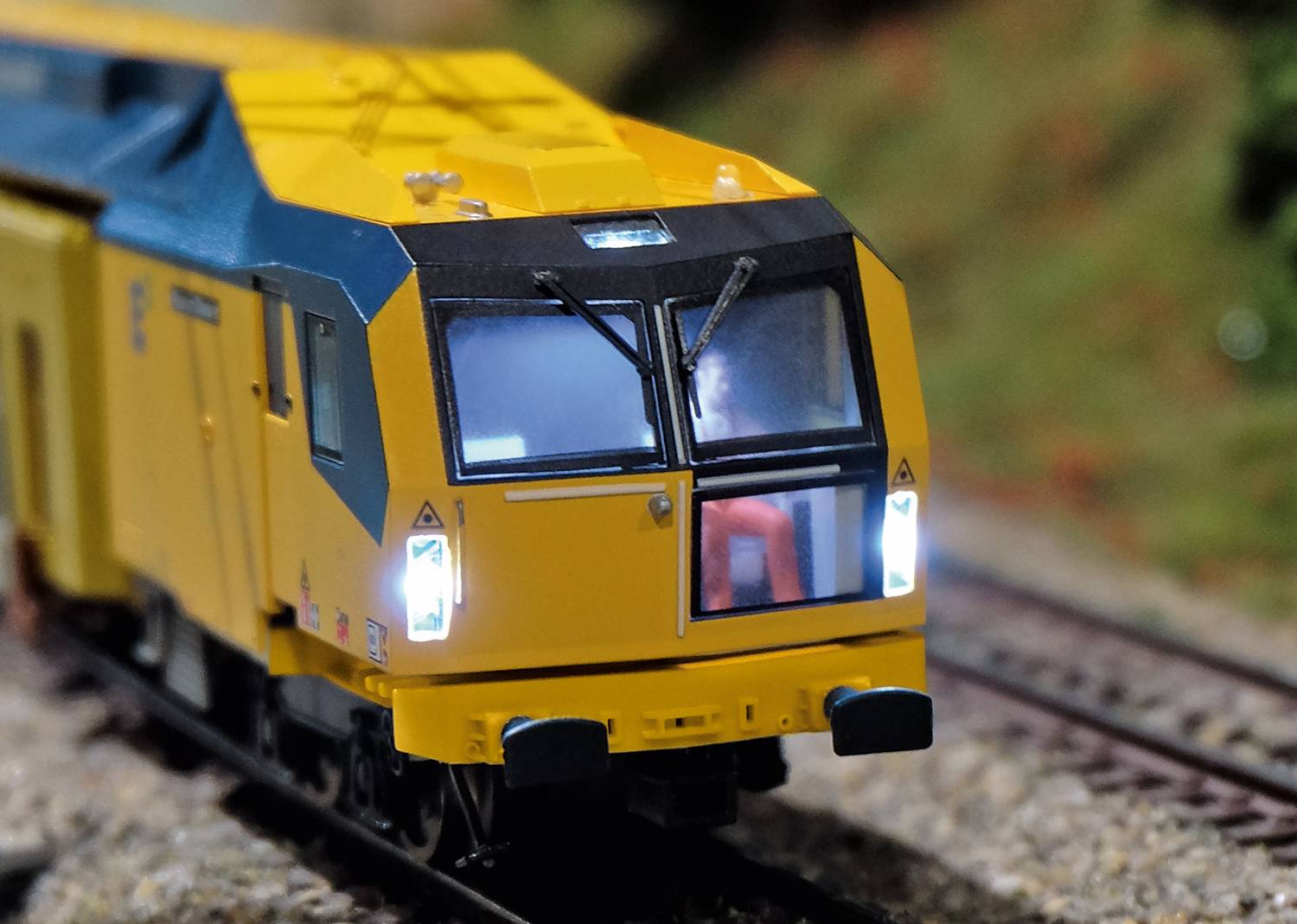 A yellow model train on a miniature railway track, front view, with lights switched on and a figure in the driver's cab.
