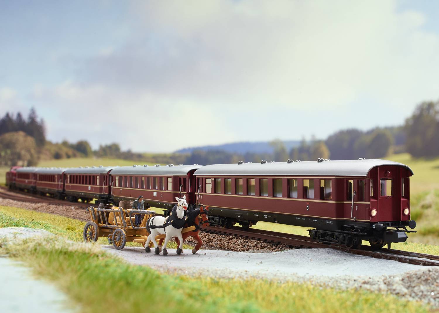 A model train travels across a railway landscape. A horse-drawn carriage is beside it on a country lane. In the background, a hilly countryside.