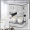 Silver espresso machine with two white coffee cups on a table. Coffee beans and another empty cup are placed beside it.