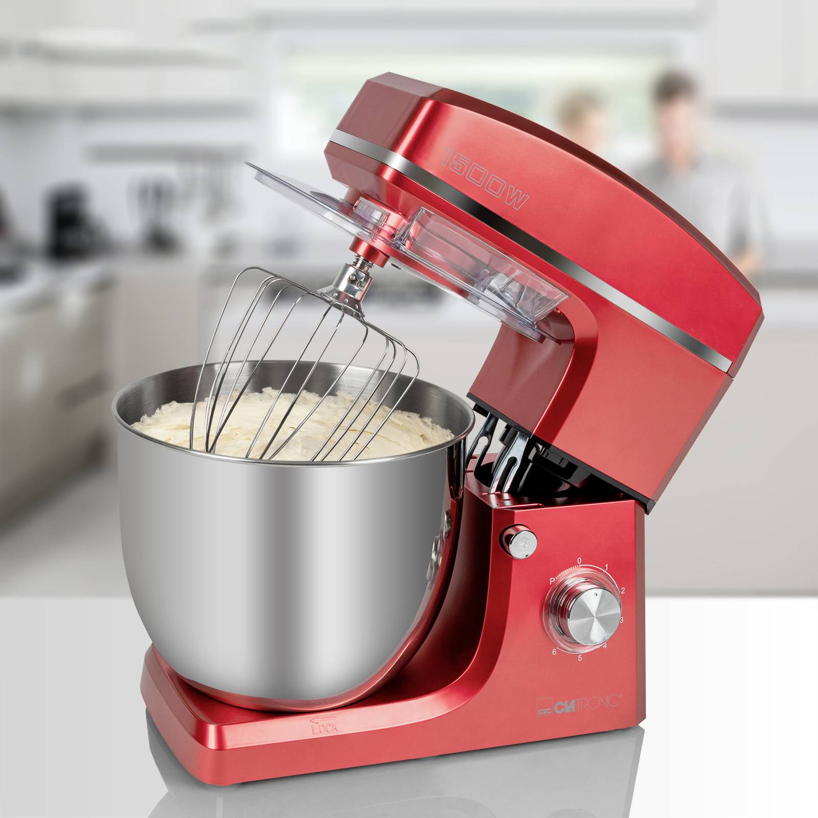 Red stand mixer with mixing bowl full of dough, set up in a modern kitchen. Two blurred people in the background.