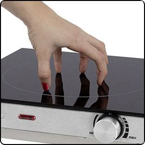 A hand carefully touches the smooth, black surface of a switched-on induction hob with a temperature control.