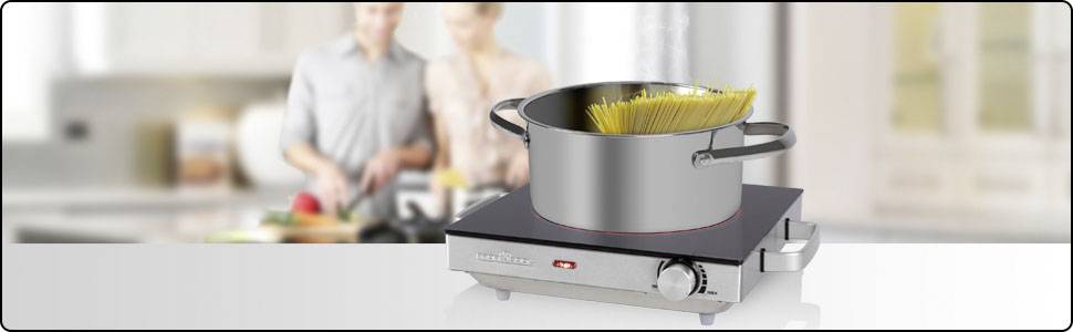 A pot of spaghetti is being cooked on an electric hob. In the background, a couple are preparing food together in the kitchen.