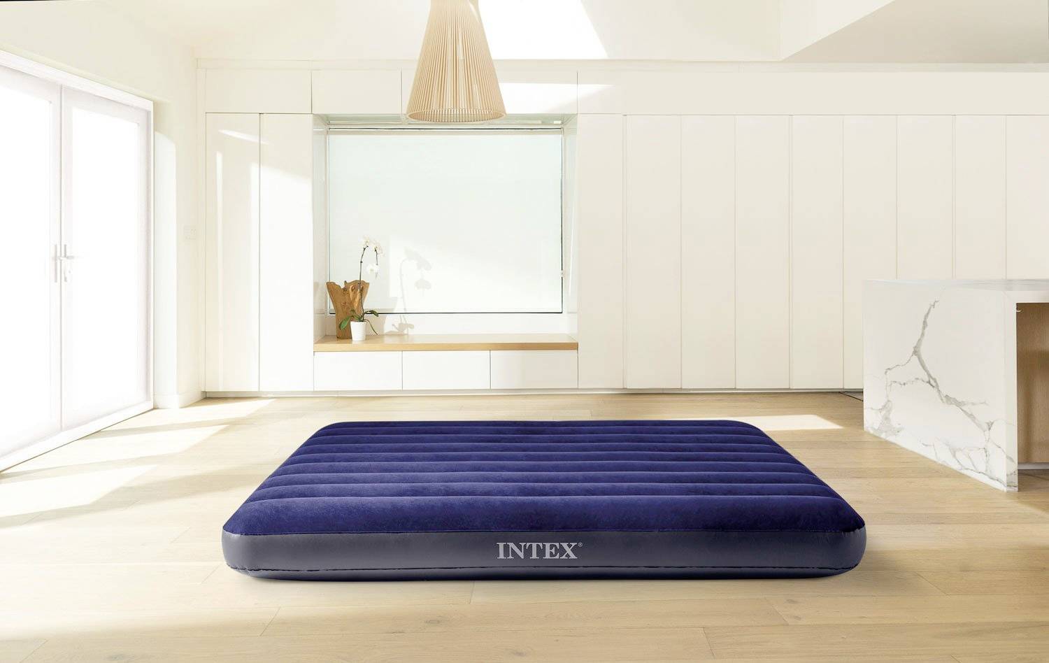 A dark blue air mattress lies on a light wooden floor in a modern, sunlit room with white walls and a window.