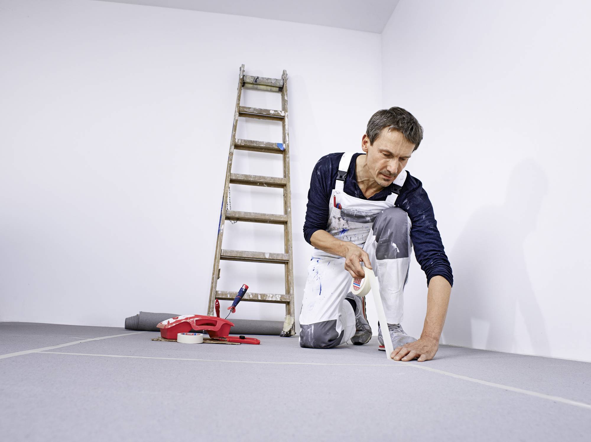 A man in workwear is laying floor tiles in a room. A ladder stands in the background, with tools lying nearby.
