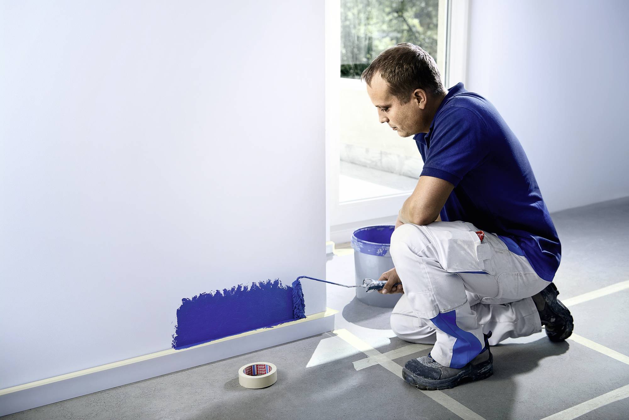 A man in workwear is painting a wall with blue paint. He is using a paint roller. A window in the background provides light.