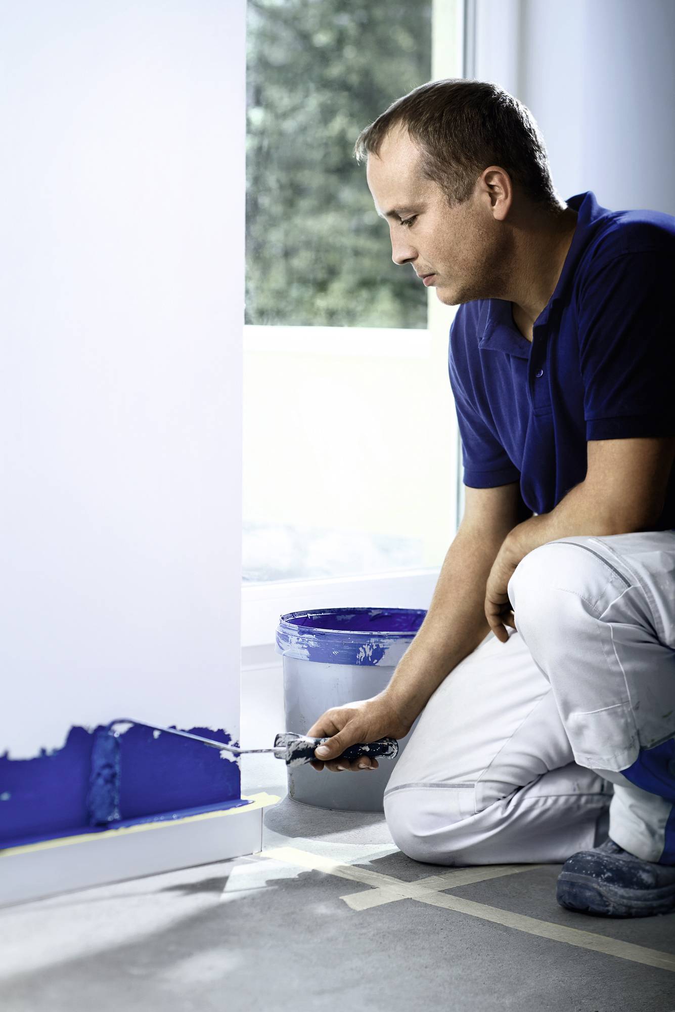 A person in blue clothing is painting a white wall with dark blue paint in a room with daylight. A paint bucket is standing nearby.