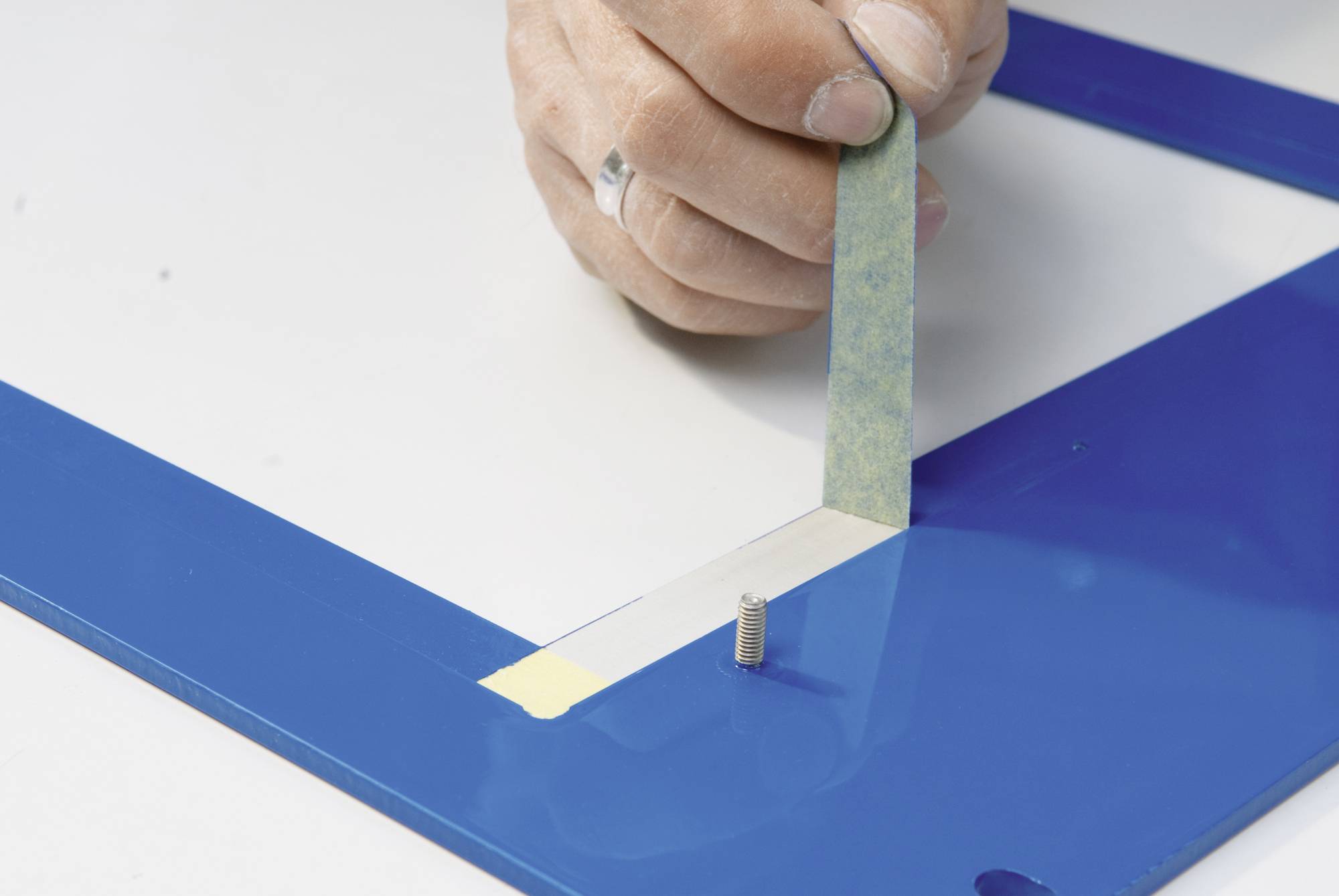 A hand carefully removes adhesive tape from a blue-painted metal frame with a screw.
