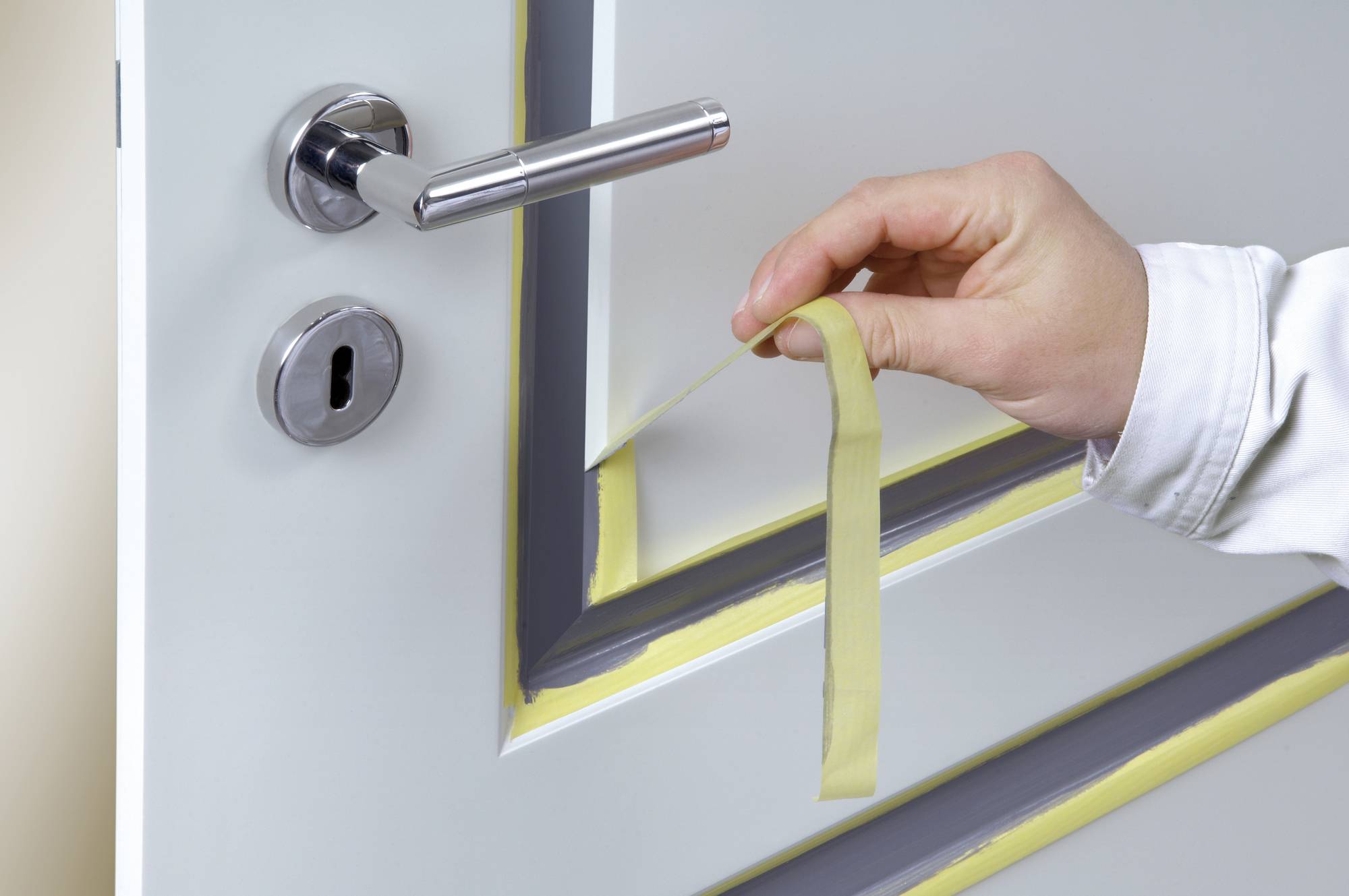 A hand is peeling yellow masking tape from a grey door. The door has a silver handle and a keyhole.