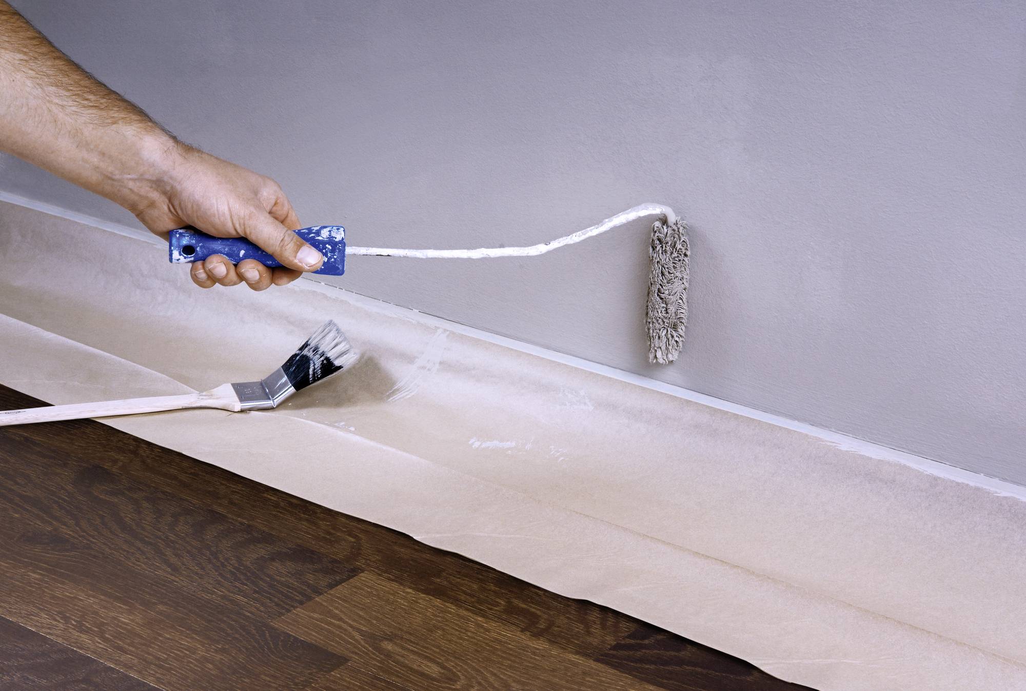 A person is painting the wall grey with a paint roller, while a paintbrush lies on the floor. The floor is covered with protective sheeting.