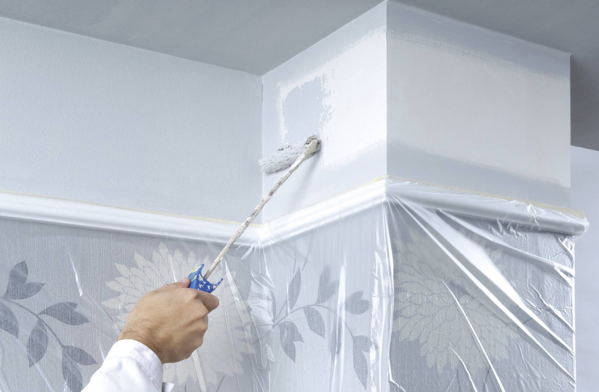 A hand with a paint roller is painting the top corner of a pale blue wall, which is covered with protective sheeting to prevent splashes.