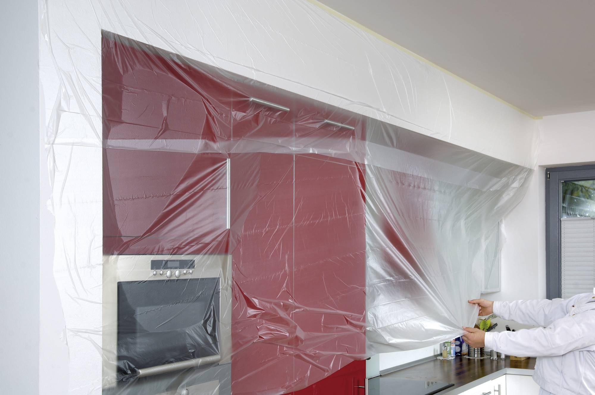 A person is covering red kitchen units and a built-in appliance with plastic sheeting, likely in preparation for painting work.