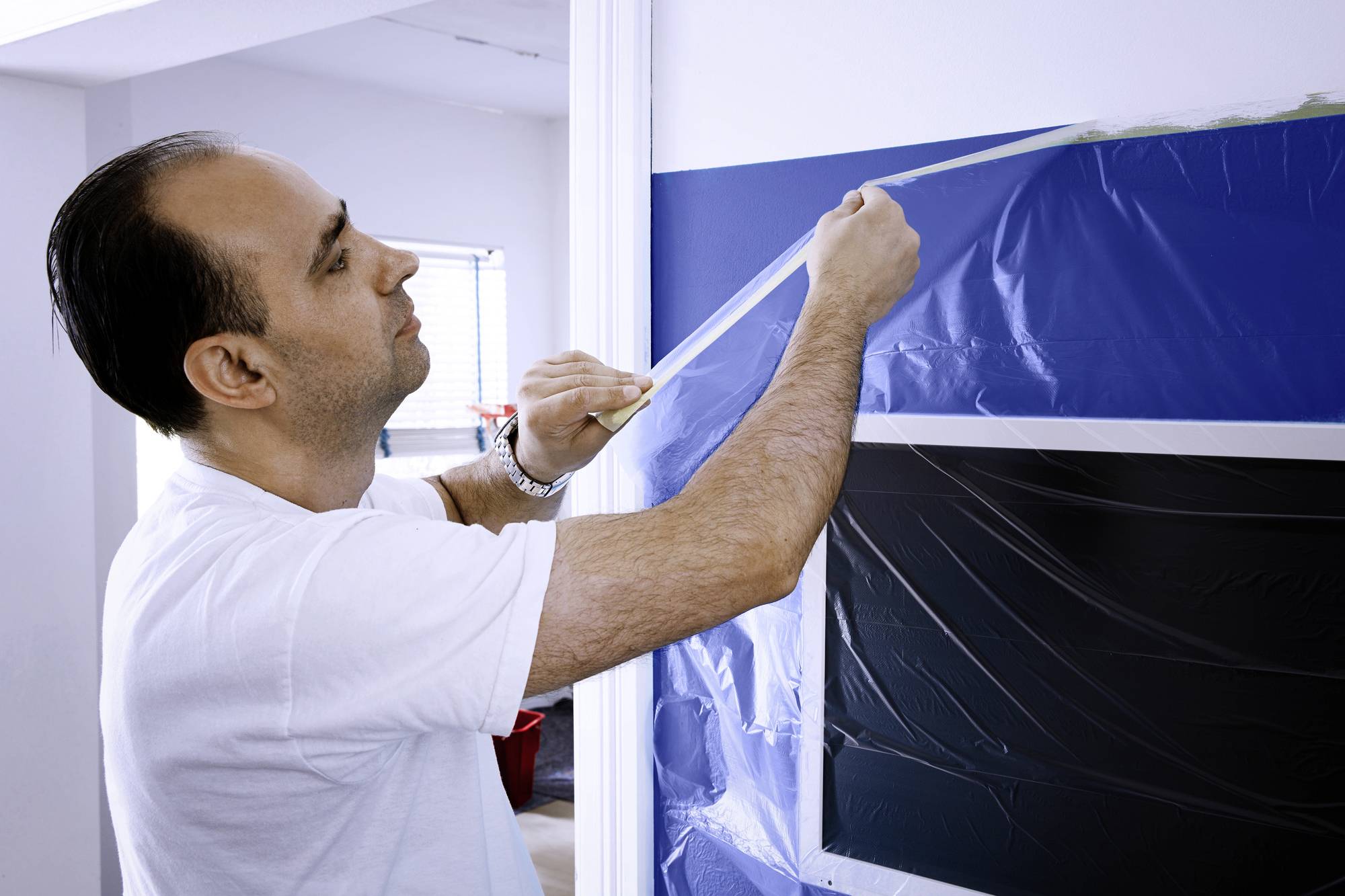 A man is applying masking tape to a wall in preparation for painting. A window can be seen in the background.