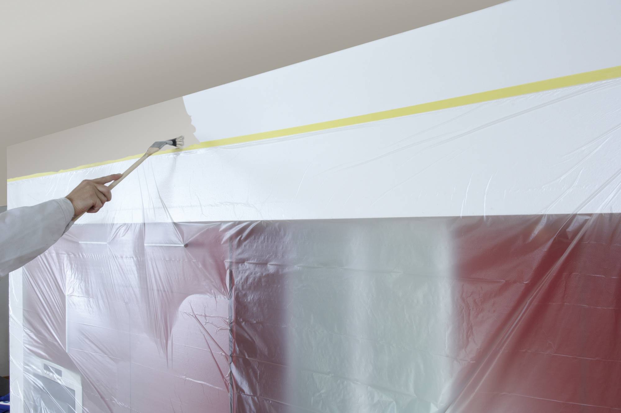 A person is painting a white wall, with part of it covered in plastic sheeting to provide protection against paint splashes.