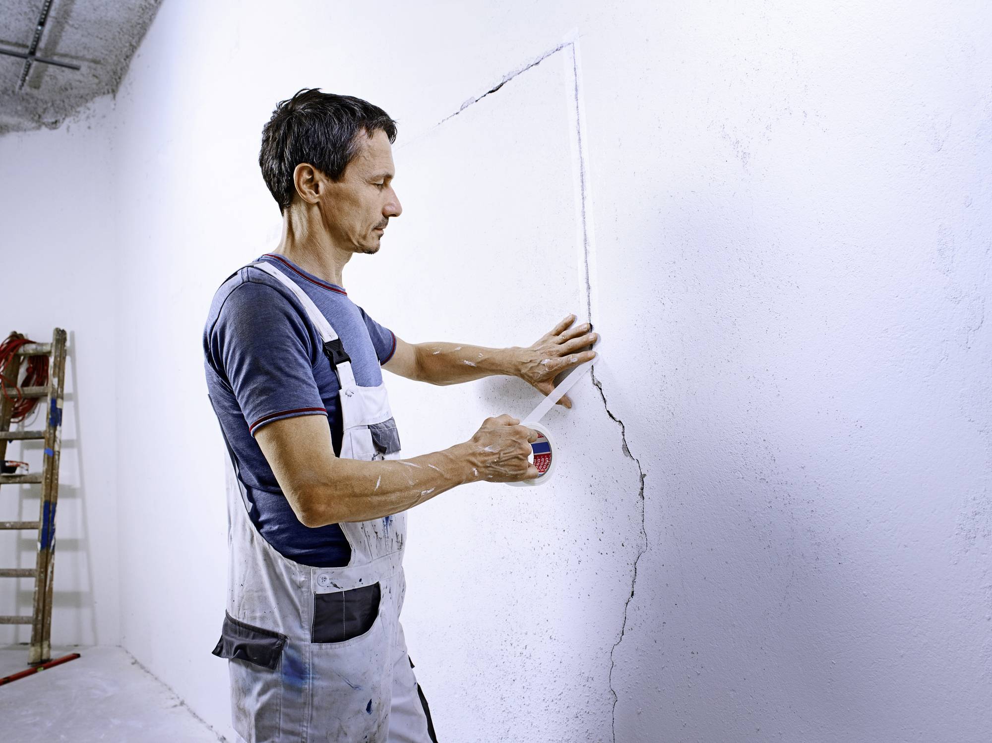 A man in workwear is repairing a crack in a wall with filler in an unfinished room. A ladder is standing in the background.
