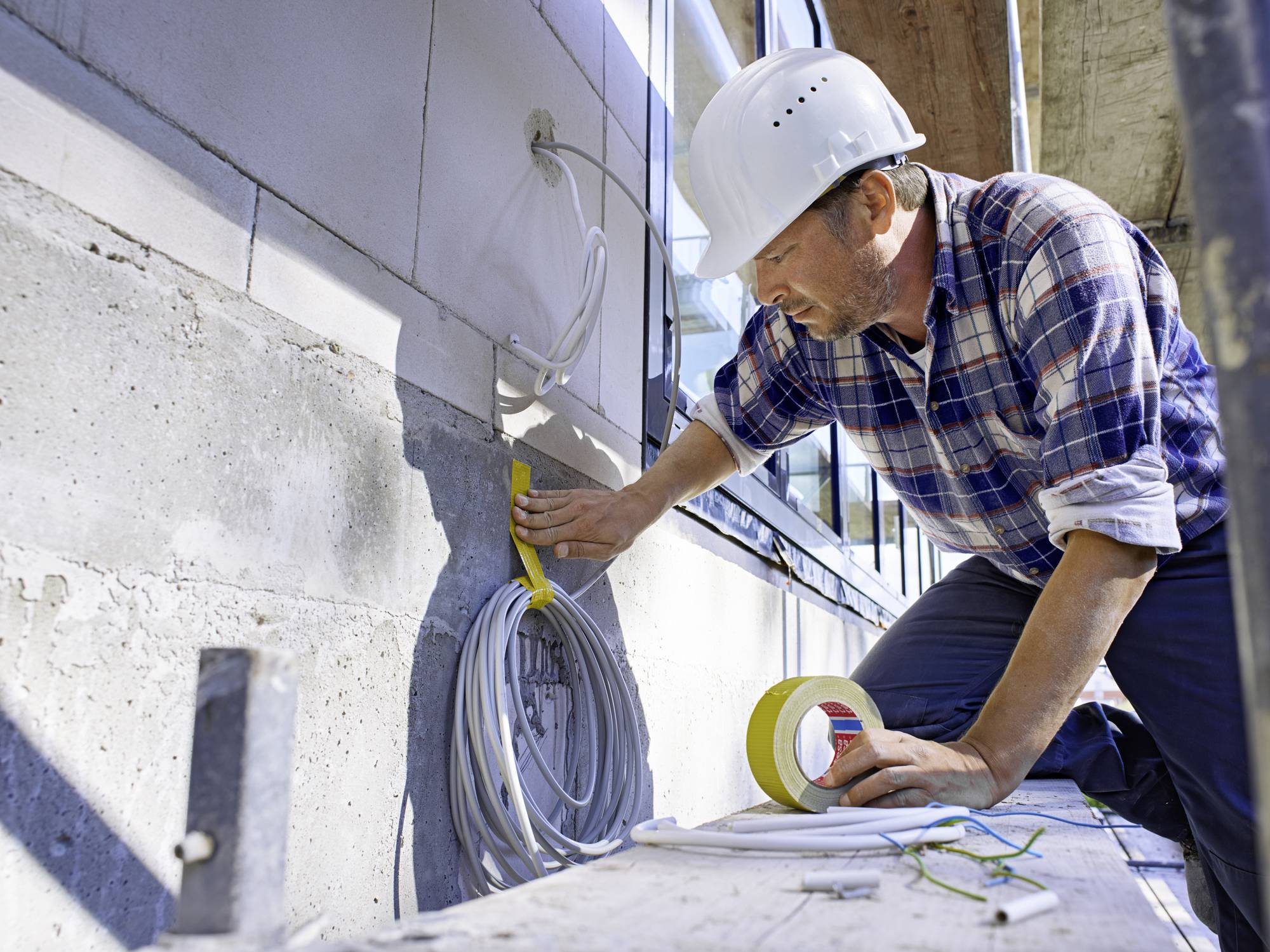 A construction worker in a checked jacket and hard hat is installing cables on a building facade and using yellow adhesive tape.
