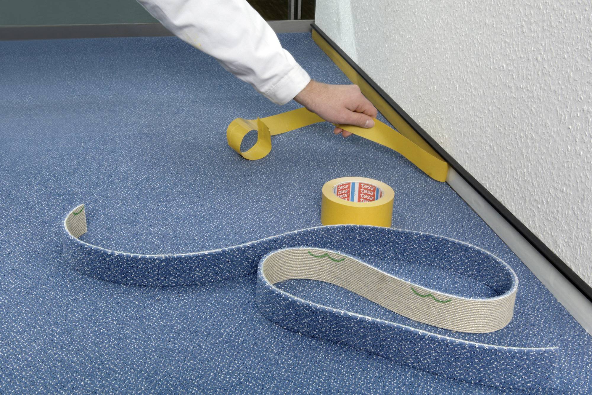 A person is sticking yellow double-sided adhesive tape along the edge of a blue fitted carpet to secure it to the wall.