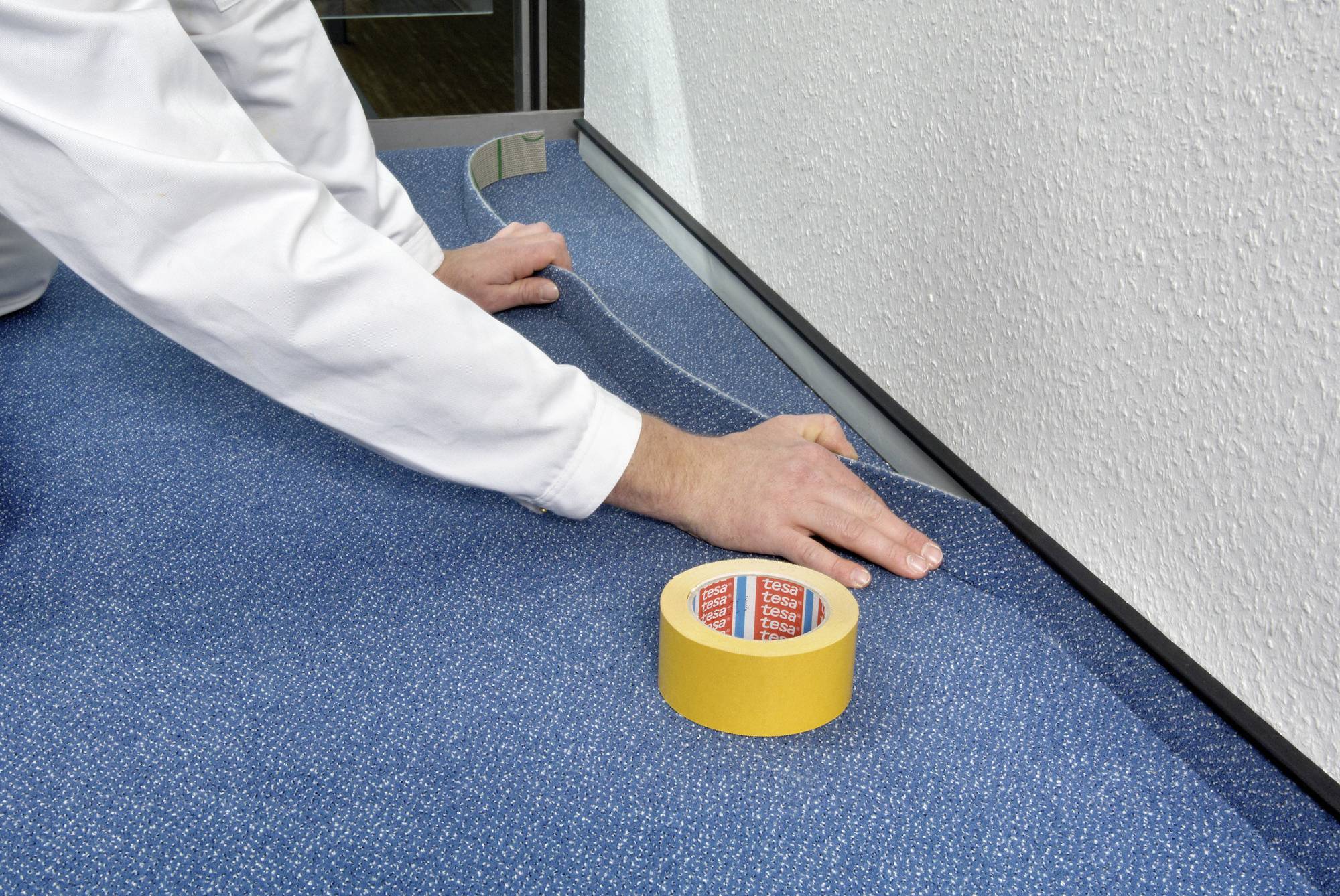 A person is laying blue carpet in a room and using yellow tape to secure the edges along the wall skirting.