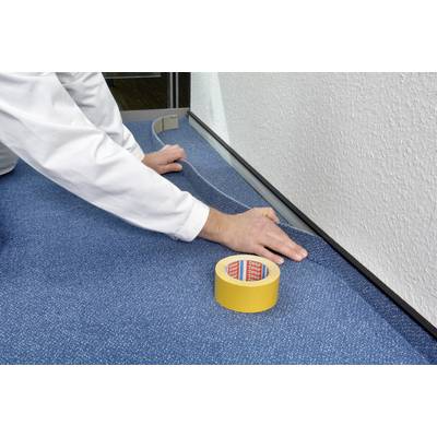 A person is laying blue carpet in a room and using yellow tape to secure the edges along the wall skirting.