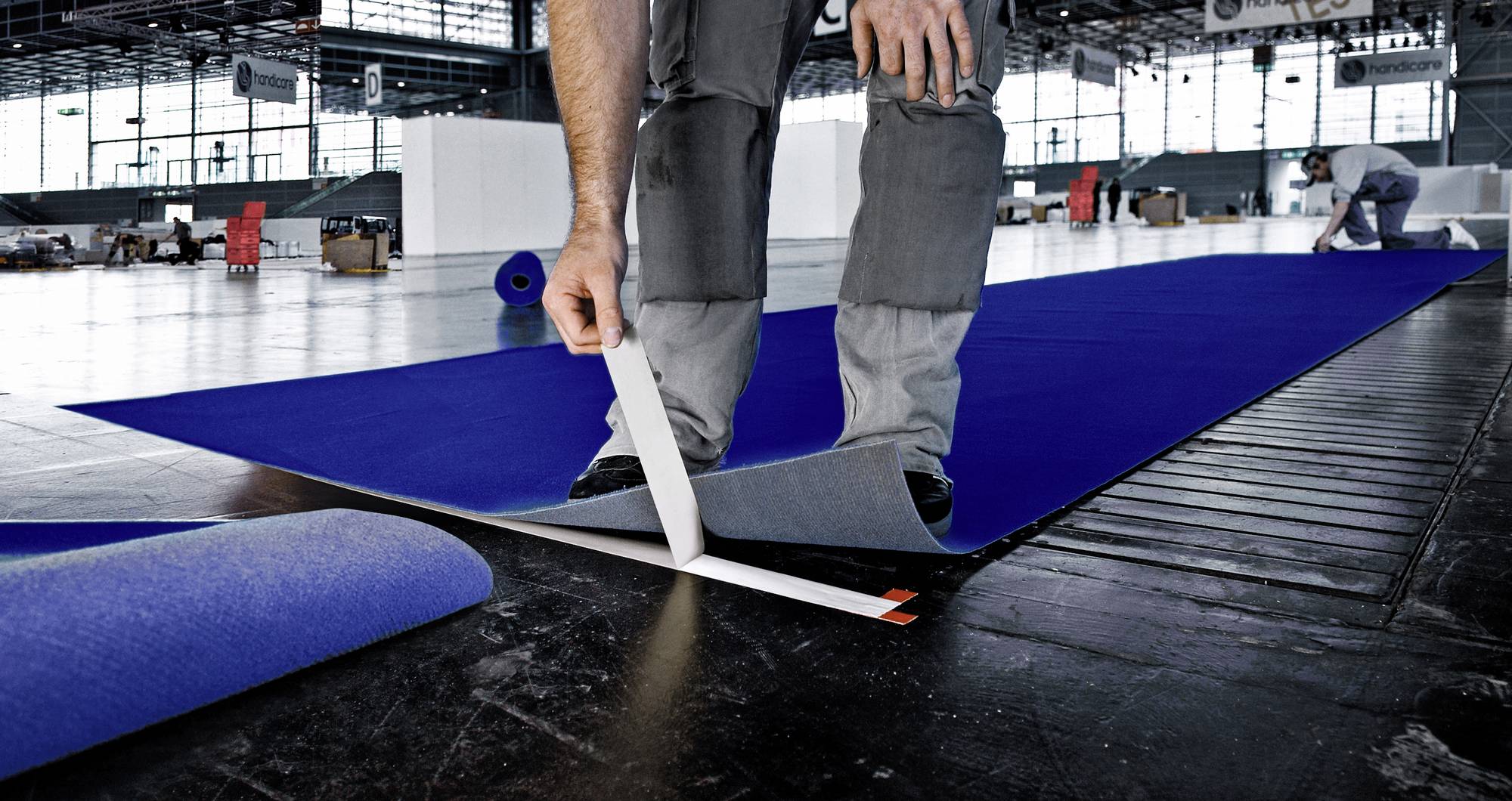 A person is laying blue carpeting in a large hall. An employee is working in the background.