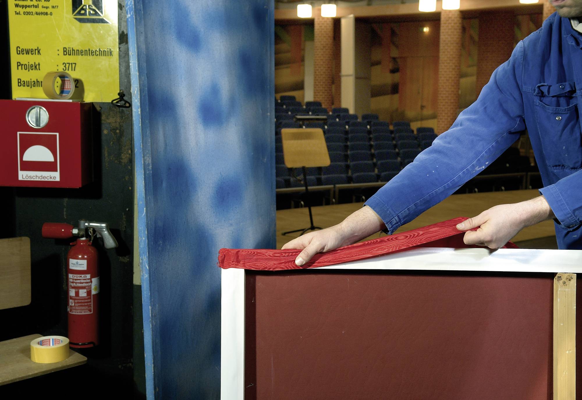 A person in blue workwear is laying red fabric on a brown surface. Empty rows of seats are visible in the background.