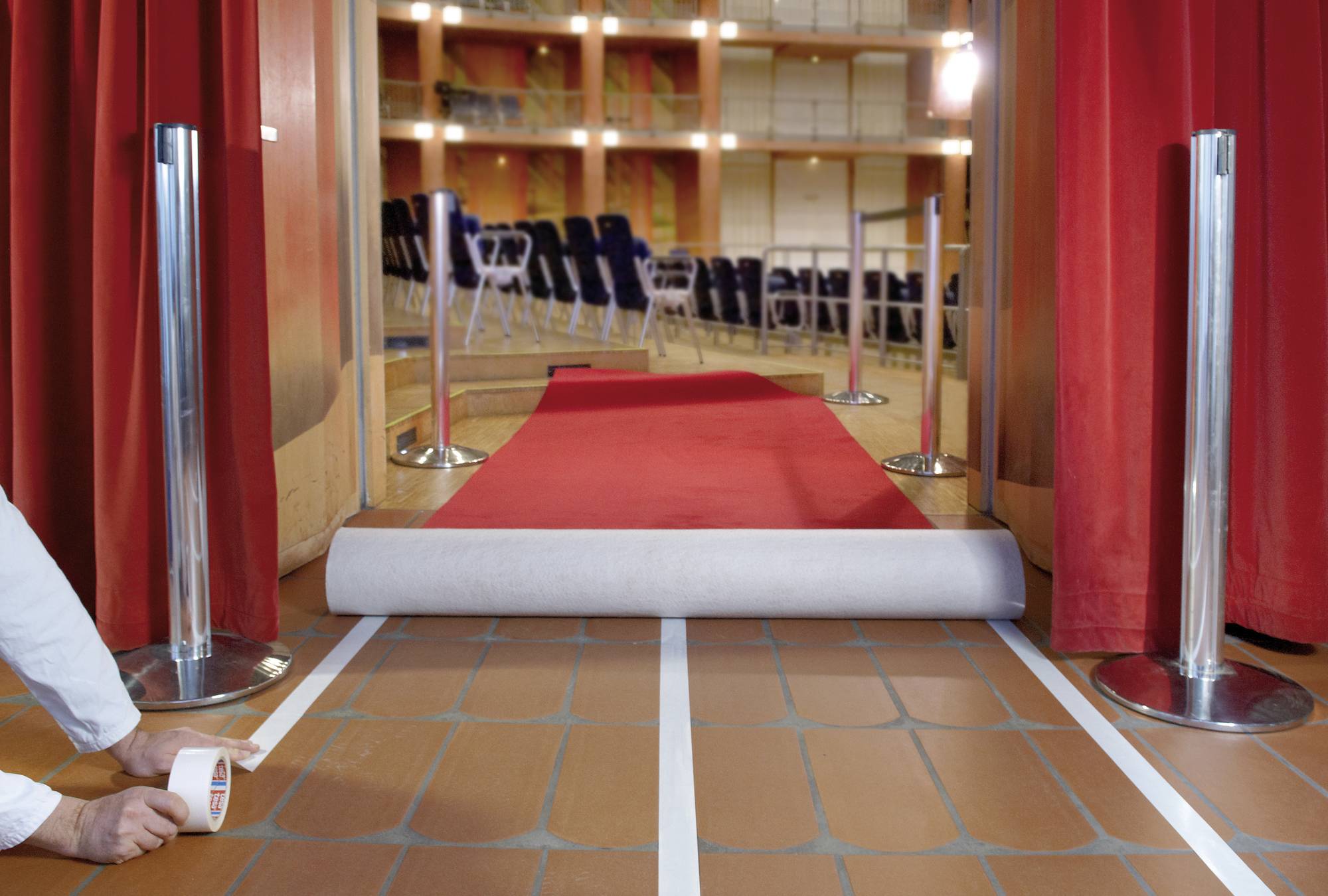 Red carpet is rolled out in a theatre auditorium, surrounded by red curtains and rows of chairs in the background.