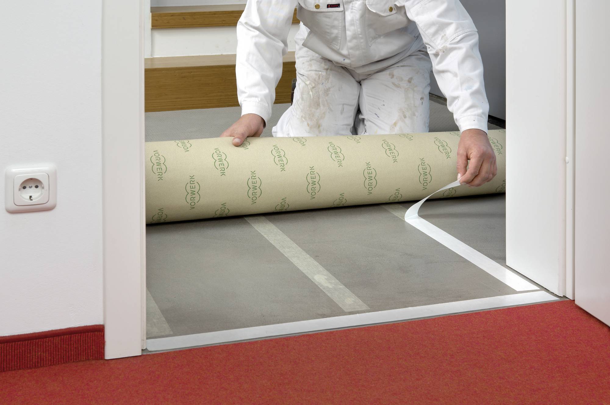 A person is laying carpet in a room with a red carpet floor by unrolling a new floor covering.