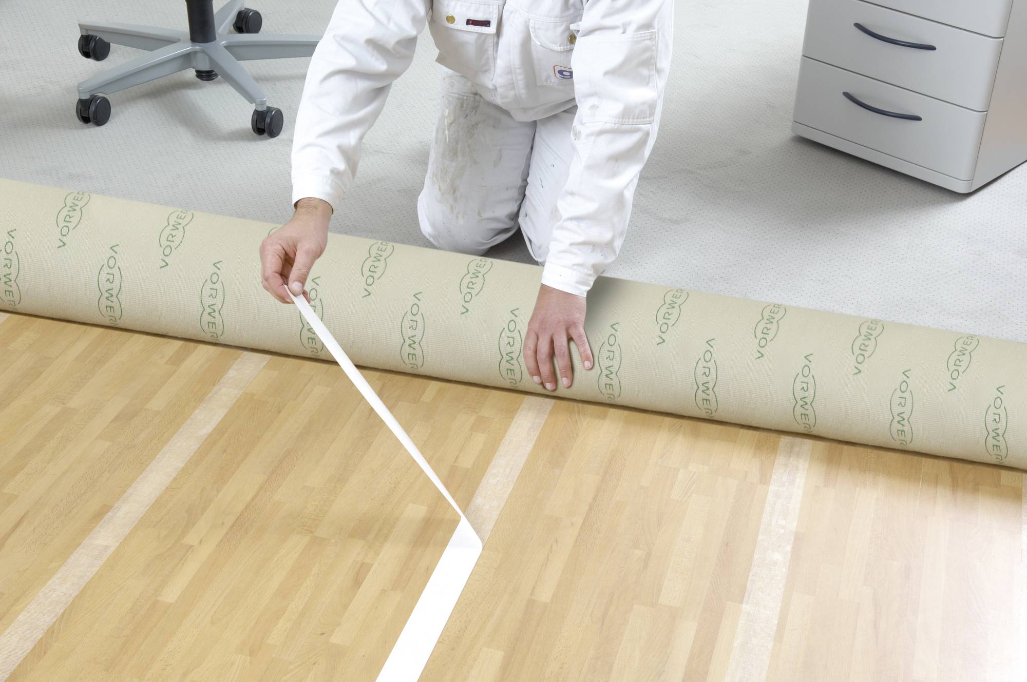 A person in workwear is laying carpet, applying tape to a wooden floor. In the background, an office chair and a cabinet can be seen.