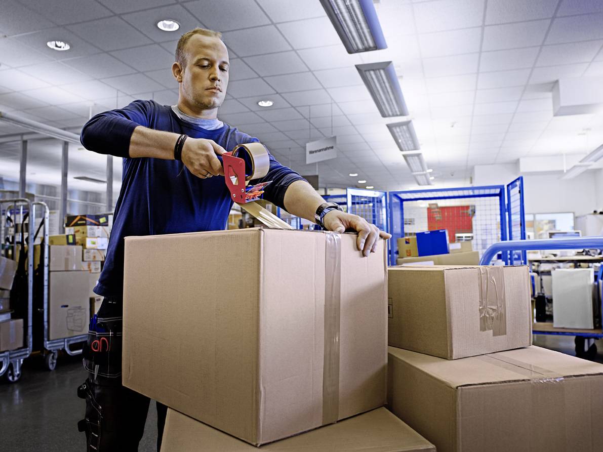 A man seals a cardboard box with tape in a storage room full of parcels, boxes, and shipping supplies.