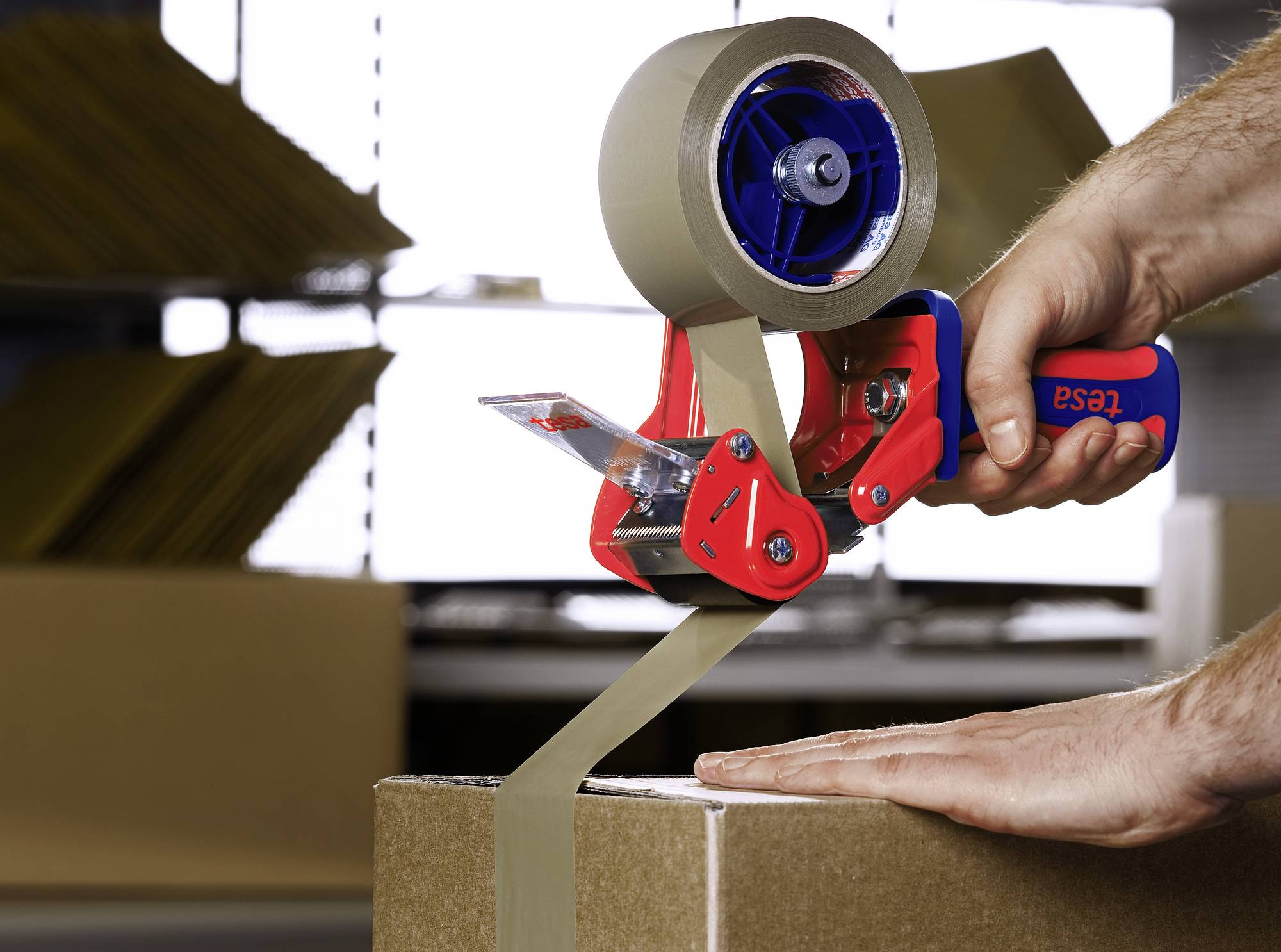 A person is using a parcel tape dispenser to seal a cardboard box. Other boxes are visible in the background.