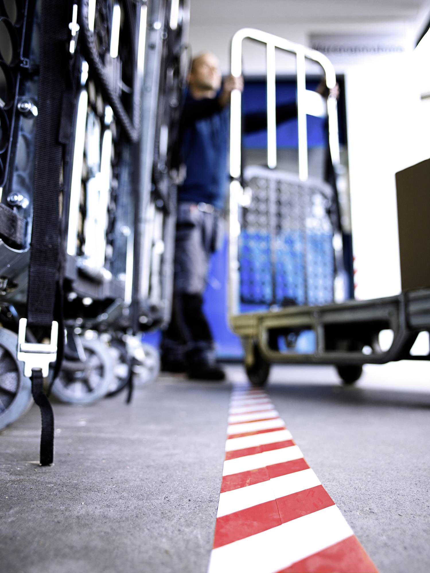 A person is pushing a trolley loaded with boxes in a storage room. A red and white striped marking delineates the floor zone.