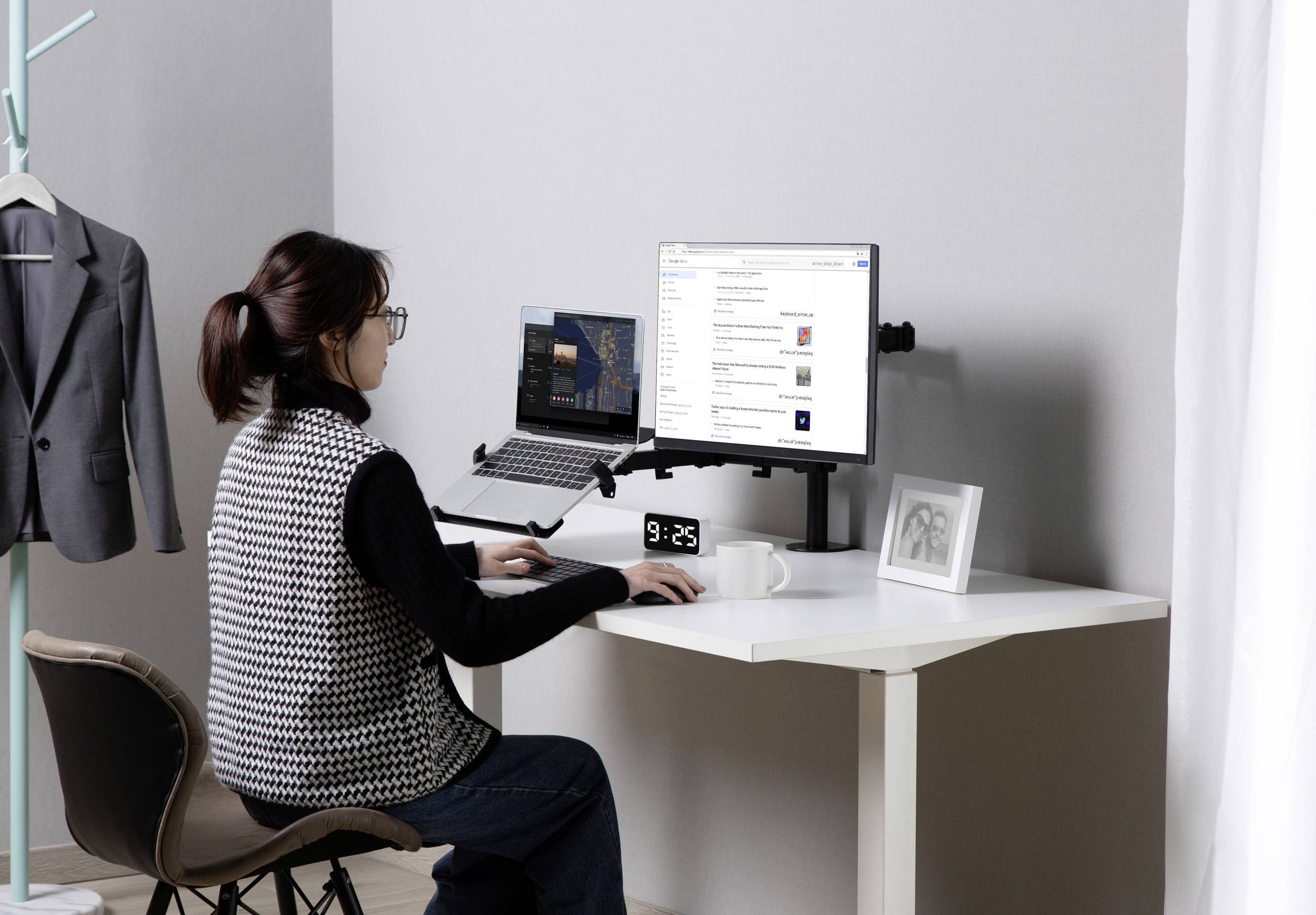 A woman is working at a desk with a laptop and monitor. The environment is modern and tidy, with a hanging blazer in the background.