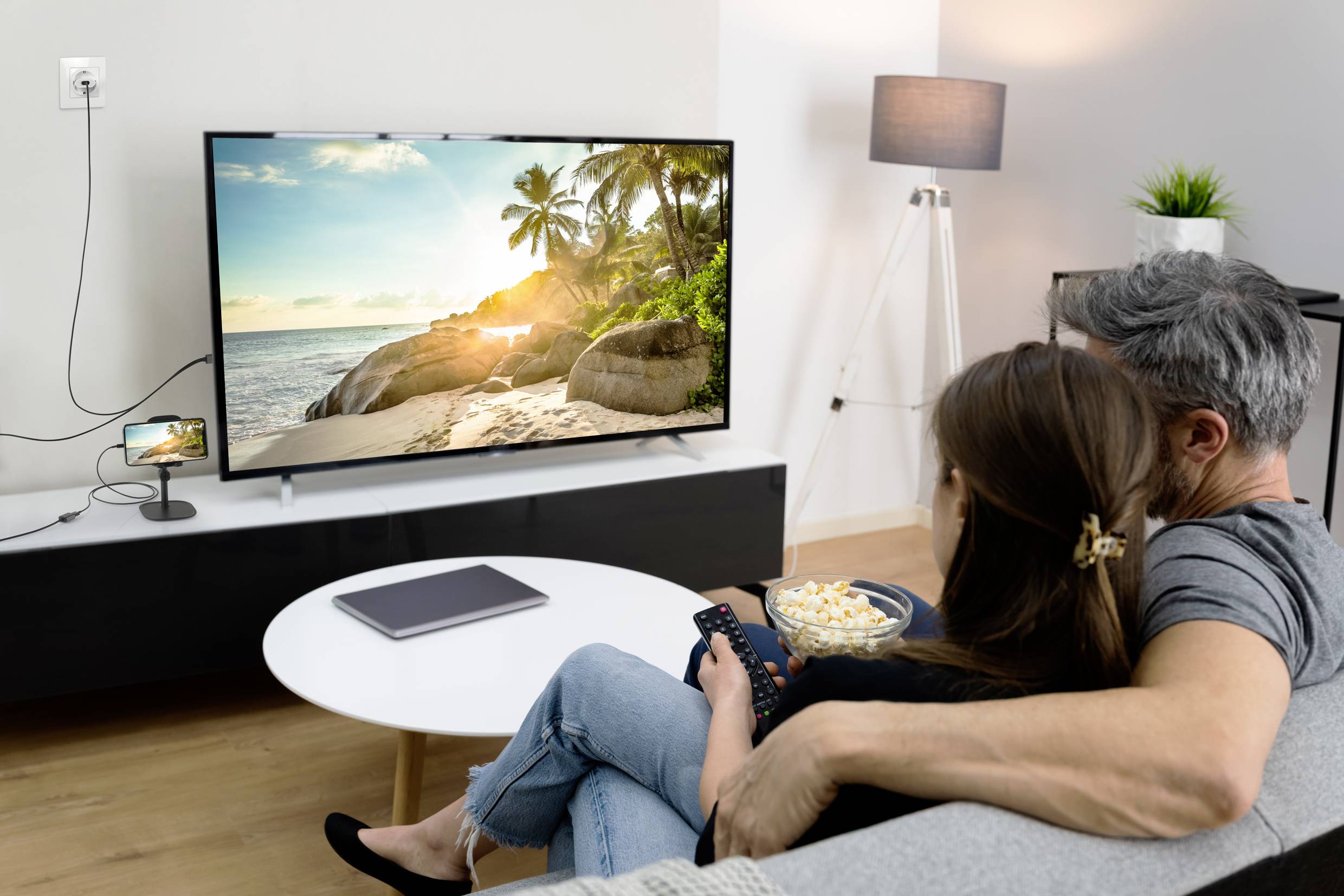 A couple are sitting on a sofa, watching a beach sunset on the television. A dining table with popcorn and a tablet are in the foreground.
