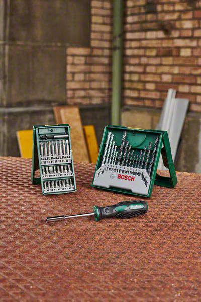 Drill set with various drill bits and a screwdriver on a metal table, with a brick wall in the background.