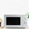 Microwave on kitchen worktop, surrounded by utensils. Egg cup and chopping board to the left, plant to the right. Minimalist kitchen design.